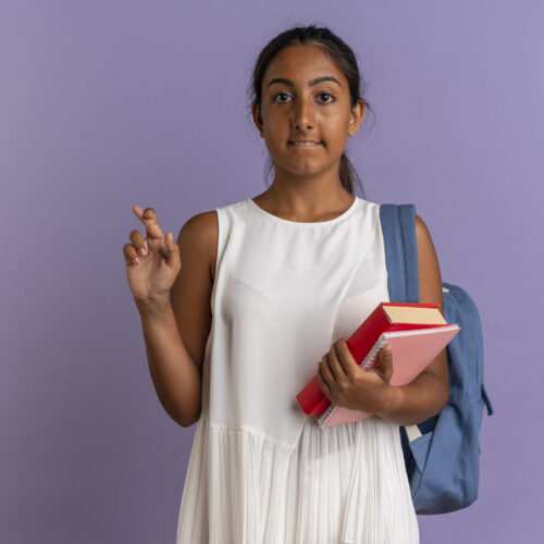 concerned young schoolgirl wearing back bag holding book with notebook and crossing fingers on isolated purple background